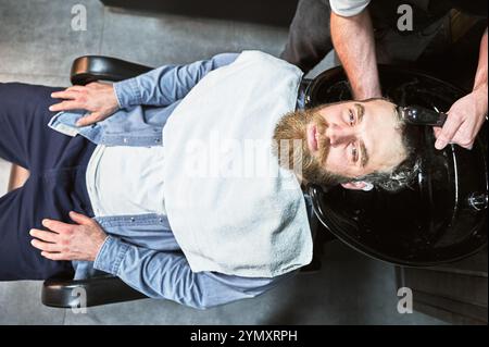 Uomo dai capelli di zenzero che si lava i capelli. Parrucchiere maschile che lava i capelli con prodotti per la pulizia profonda. Bel cliente che si rilassa con un asciugamano sul petto. Foto Stock