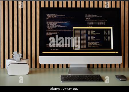 Computer desktop moderno che visualizza linee di codifica con sfondo in legno e visore per la realtà virtuale posizionato sul tavolo accanto alla tastiera Foto Stock