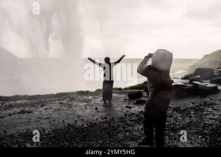 Dietro la maestosa cascata islandese: I turisti che catturano la bellezza di Seljalandsfoss si stagliano contro la nebbia e lo spruzzo in un iconico Natur islandese Foto Stock