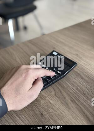 Una mano che interagisce con una calcolatrice nera su una scrivania di legno in un ufficio Foto Stock