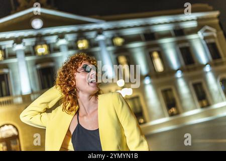 Donna sicura di sé con abiti e occhiali da sole gialli informali che si godono la notte da sola in città Foto Stock