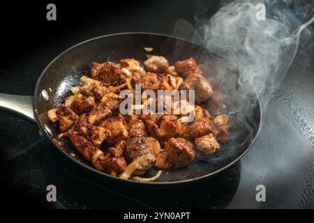 Il maiale a dadini con cipolle e condimento con paprika viene arrostito con abbondante vapore in una padella di ghisa su un piano cottura a induzione nero, cucinando un piatto di carne come il gou Foto Stock