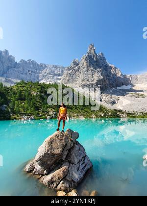 Sotto un cielo azzurro luminoso, un individuo posa con sicurezza su una roccia, circondato da acque turchesi mozzafiato e vette torreggianti, il Lago di Sorapis Dolomiti Italia Foto Stock