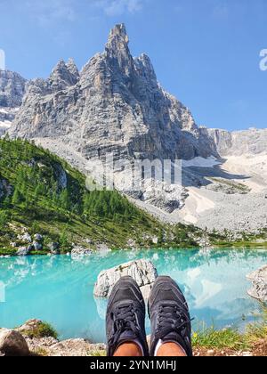 Un paio di piedi riposa accanto a un lago turchese mozzafiato, circondato da torreggianti montagne sotto un cielo azzurro cristallino, il Lago di Sorapis Dolomiti Italia Foto Stock