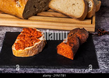 Fetta di pane con sobrasada maiorchina e accanto a un budello di salsiccia su un piatto di pietra nera, con una pagnotta tagliata a fette nel dorso Foto Stock