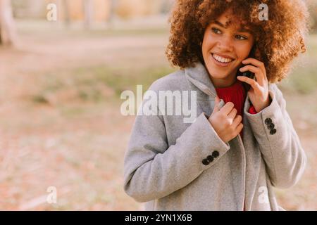 Una giovane donna con i capelli ricci sorride brillantemente mentre parla al telefono, vestita calorosamente con un cappotto elegante in mezzo a un pittoresco paesaggio autunnale Foto Stock