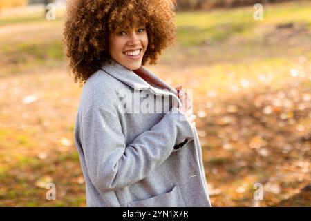 La brillante giornata autunnale affascina mentre una giovane donna cammina attraverso un incantevole parco, vestito con un comodo cappotto grigio e un maglione rosso, che incarna calore e gioia in mezzo Foto Stock