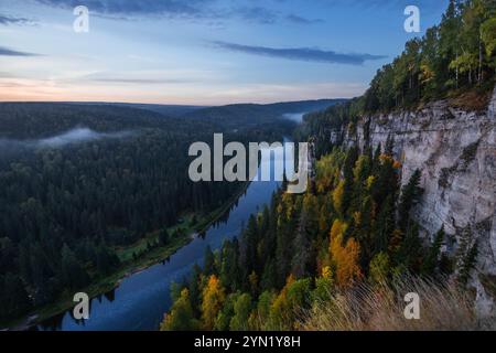 Un tranquillo fiume scorre dolcemente attraverso lussureggianti foreste verdi e torreggianti scogliere al crepuscolo, con la nebbia che si innalza sopra l'acqua e il vibrante fogliame autunnale illu Foto Stock