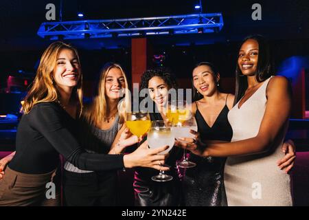 Foto di gruppo di amici gioiosi che sorseggiano cocktail e sorridono brillantemente in un vivace nightclub. Stanno ridendo e brindando con cocktail colorati, Foto Stock