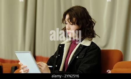 Una ragazza premurosa si appoggia sul suo notebook, immersa nella scrittura mentre è seduta su una sedia in stile teatro. Questa immagine trasmette temi di determinazione Foto Stock