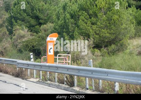 Telefono stradale di emergenza arancione con segnale sos e numero 825 situato su un'autostrada con guardrail e foresta sullo sfondo Foto Stock