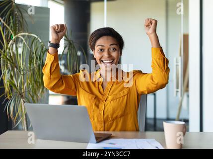 Successo aziendale. Eccitata giovane donna d'affari nera che gestiva sì, scuotendo pugni di gioia, seduta a un tavolo con un notebook Foto Stock