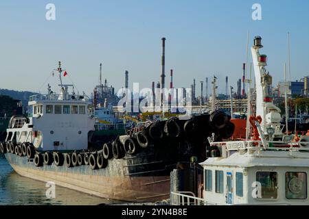 Ulsan, Corea del Sud - 14 novembre 2024: Vista ravvicinata delle barche attraccate al porto di Jangsaengpo, incorniciate da camini torreggianti e impianti industriali Foto Stock