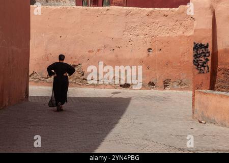 In un giorno d'estate molto caldo, una donna solitaria si trascina sul lato ombreggiato di una strada surriscaldata nella medina di Marrakech. Foto Stock