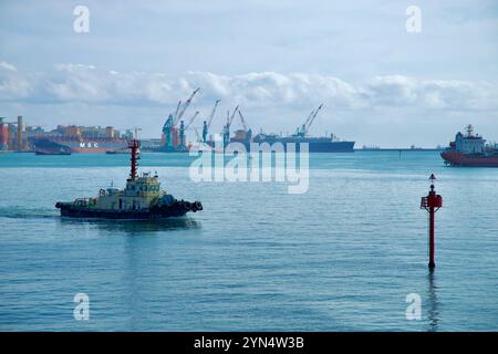 Ulsan, Corea del Sud - 14 novembre 2024: Un rimorchiatore naviga attraverso le acque del porto di Ulsan, con navi portacontainer, gru industriali e marittime Foto Stock