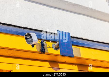 Telecamera di sicurezza per esterni montata su una parete gialla con pannello solare, che cattura la luce solare per una sorveglianza ecocompatibile. Foto Stock