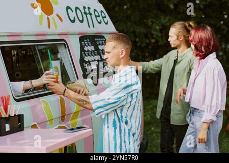 Vista laterale dell'uomo caucasico che prende la limonata e l'hot dog mentre gli altri clienti leggono il menu sul camion del cibo Foto Stock