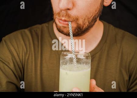 Un bicchiere trasparente con una bevanda e una cannuccia da cui un uomo beve. La foto mostra un uomo che beve un latte attraverso una cannuccia Foto Stock