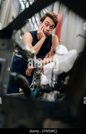 Meccanico dubbio che ispeziona il motore dell'auto in officina, perplesso e frustrato, mostrando le sfide della manutenzione automobilistica. Foto Stock
