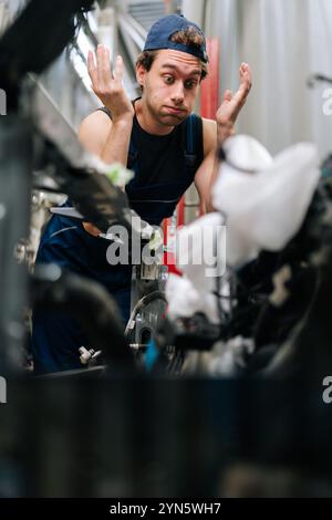 Ritratto verticale di un riparatore confuso sconcertato durante l'ispezione del motore di un'auto in riparazione. Maschio meccanico che guarda il motore dell'auto e gesti con Foto Stock