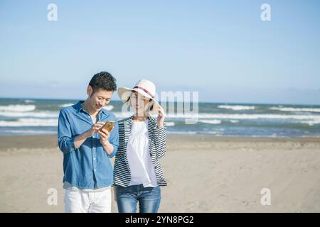 Coppia di 20 e 30 anni che cammina su una spiaggia mentre guarda il telefono Foto Stock