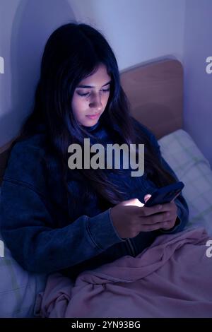 Immagine verticale di adolescente triste e stanca che utilizza uno smartphone sdraiato a letto a tarda notte, che gioca, guarda video e digita. Sc. Bambini Foto Stock
