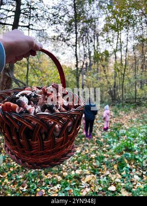 Un cesto pieno di funghi appena raccolti si trova in una vibrante foresta autunnale. Sullo sfondo, una famiglia cammina attraverso le foglie cadute, godendosi Foto Stock