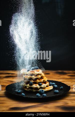 Waffle belgi con mirtilli, cosparsi di zucchero a velo. Waffle con frutti di bosco su sfondo nero su un tavolo di legno. Foto di alta qualità Foto Stock