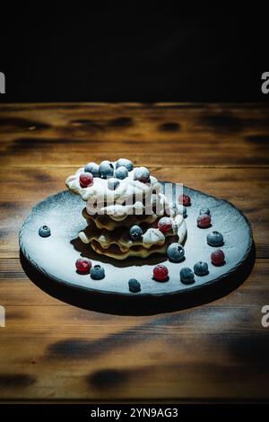 Waffle belgi con mirtilli rossi e mirtilli cosparsi di zucchero a velo su un tavolo di legno su un piatto nero. Foto di alta qualità Foto Stock