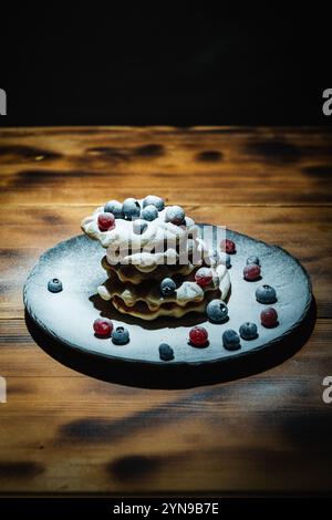 Waffle belgi con mirtilli rossi e mirtilli cosparsi di zucchero a velo su un tavolo di legno su un piatto nero. Foto di alta qualità Foto Stock