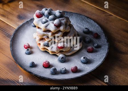 Waffle belgi con mirtilli rossi e mirtilli cosparsi di zucchero a velo su un tavolo di legno su un piatto nero. Foto di alta qualità Foto Stock