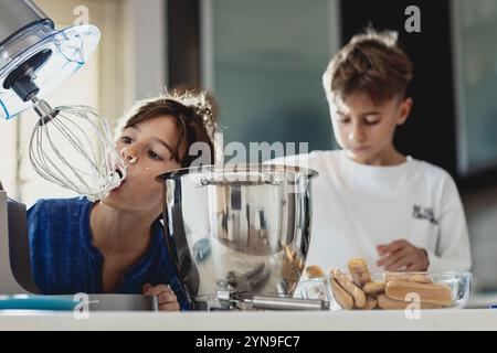 Due bambini potranno cucinare in una cucina moderna. Un bambino assaggia gli ingredienti di un mixer mentre l'altro prepara gli ingredienti per un dessert. Il Foto Stock