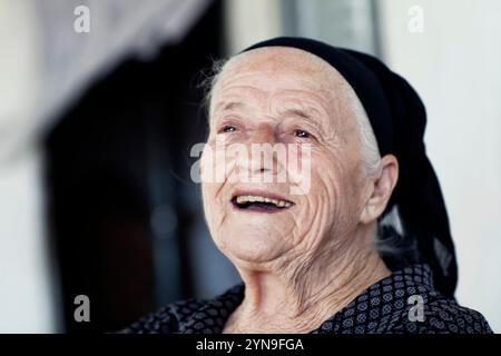 Una donna anziana con un sorriso caldo siede comodamente in casa, indossando una sciarpa nera sulla testa e un abito a fantasia. La sua espressione irradia gioia A. Foto Stock