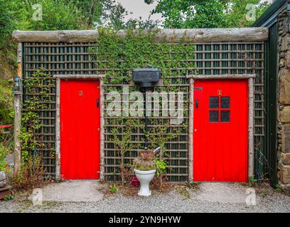 Una toilette rustica presenta due porte rosse incorniciate da alberi di legno adornati da piante rampicanti. Il verde lussureggiante circonda il caratteristico struc Foto Stock