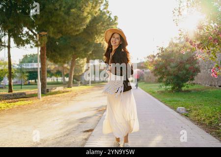 Una bella donna vestita con un abito bianco passeggiando per un parco durante l'ora d'oro Foto Stock