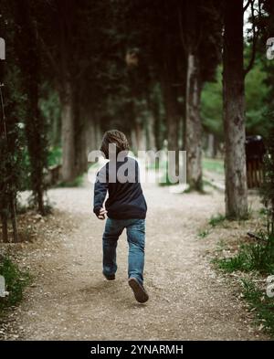 Un bambino corre gioiosamente lungo un sentiero di ghiaia circondato da alberi alti in un parco. La luce del sole filtra attraverso le foglie, creando un'atmosfera tranquilla Foto Stock