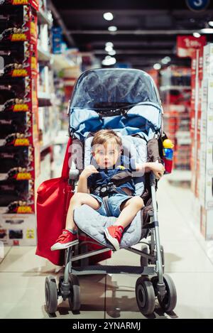 Un bambino piccolo occupa un passeggino in un corridoio del supermercato circondato da scaffali riforniti di vari prodotti. Il bambino sembra rilassato mentre fa shopping Foto Stock