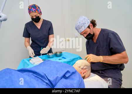 Due chirurghi in una clinica che preparano ed eseguono una procedura di trapianto di capelli su un paziente utilizzando strumenti medici e anestesia. Foto Stock