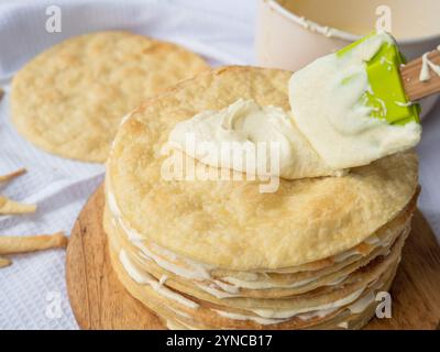 Spatola in silicone che distribuisce la crema alla vaniglia su una pila di sottili strati di burro rotondi cotti su un asse di legno, una ciotola e un nuovo pelo che ti aspettano sul retro Foto Stock