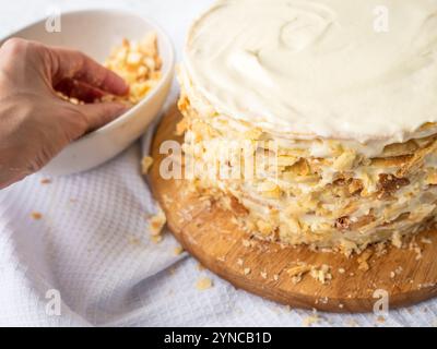 Una spolverata a mano che decora le briciole su una pila di sottili strati rotondi spalmati con crema alla vaniglia, una ciotola di impasto su un asciugamano bianco per waffle. Foto Stock