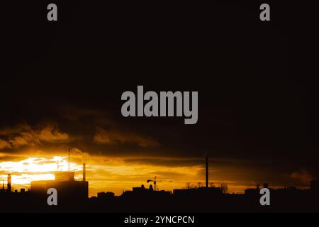 La silhouette di una città industriale con i suoi camini che fumano durante un tramonto luminoso contro un cielo scuro e nuvoloso causato dalle emissioni. Questa è una res Foto Stock