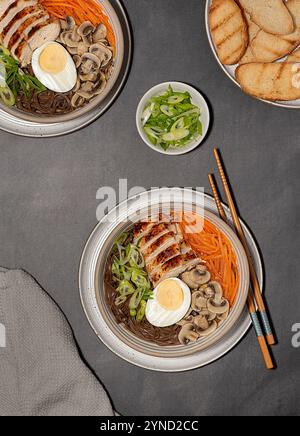 Fotografia gastronomica di zuppa di ramen con pollo, soba, spaghetti di grano saraceno, uova, funghi, cipolla primaverile e carote, toast, bacchette Foto Stock