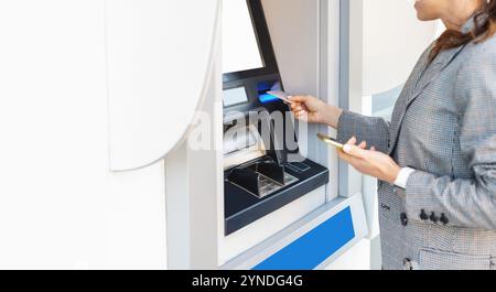 Donna d'affari in un elegante outfit casual che utilizza un bancomat con smartphone e carta di credito sulla strada cittadina Foto Stock