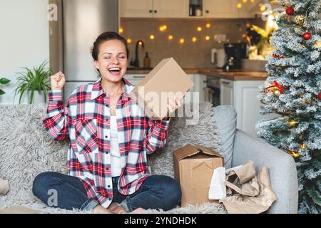 Donna caucasica felice in camicia a quadri seduto sul divano di casa con scatole di cartone che celebrano l'acquisto online e ricevono la consegna. Foto Stock