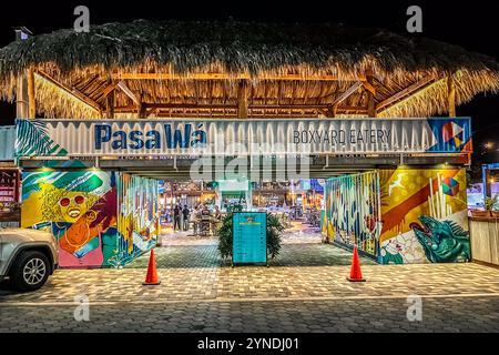Il Pasawá Box Eatery si trova su Caracasbaaiweg a Willemstad, Curaçao, qui raffigurato di notte: il locale di street food, ospitato in container navali riconvertiti, ha un tetto in foglie di palma e murales colorati. La decorazione della facciata è opera dell’artista di Amsterdam Sander van Beusekom (nome d’arte BLEND), che ha anche creato il murale storico sull’acqua su John F. Kennedy Boulevard. I visitatori si servono da soli a vari stand di cibo all’interno del locale. Foto Stock