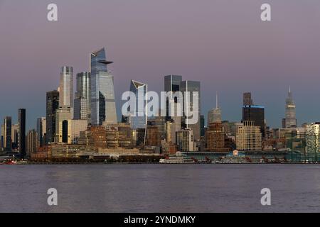 Lo skyline di Midtown di New York si illumina di sfumature del tramonto riflesse su eleganti edifici moderni, mentre il fiume Hudson si estende serenamente in primo piano. Foto Stock