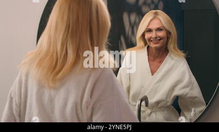 Caucasico bella donna anziana matura donna di mezza età in bagno signora in accappatoio guardare specchio felice sorriso mattina relax salute benessere Foto Stock