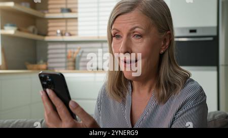 Sorpreso felice anziano 60s donna caucasica leggere buone notizie utilizzando il telefono cellulare sul divano a casa il preavviso bancario ha stupito la pensionessa donna che vince online Foto Stock