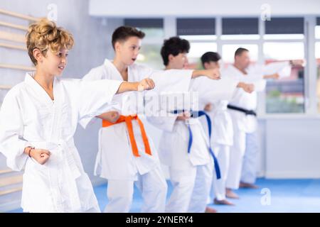 Bambini in kimono in fila sul tatami e che praticano colpi in palestra Foto Stock