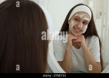 Adolescente con schiuma detergente sul viso vicino allo specchio in casa. Trattamento dell'acne Foto Stock